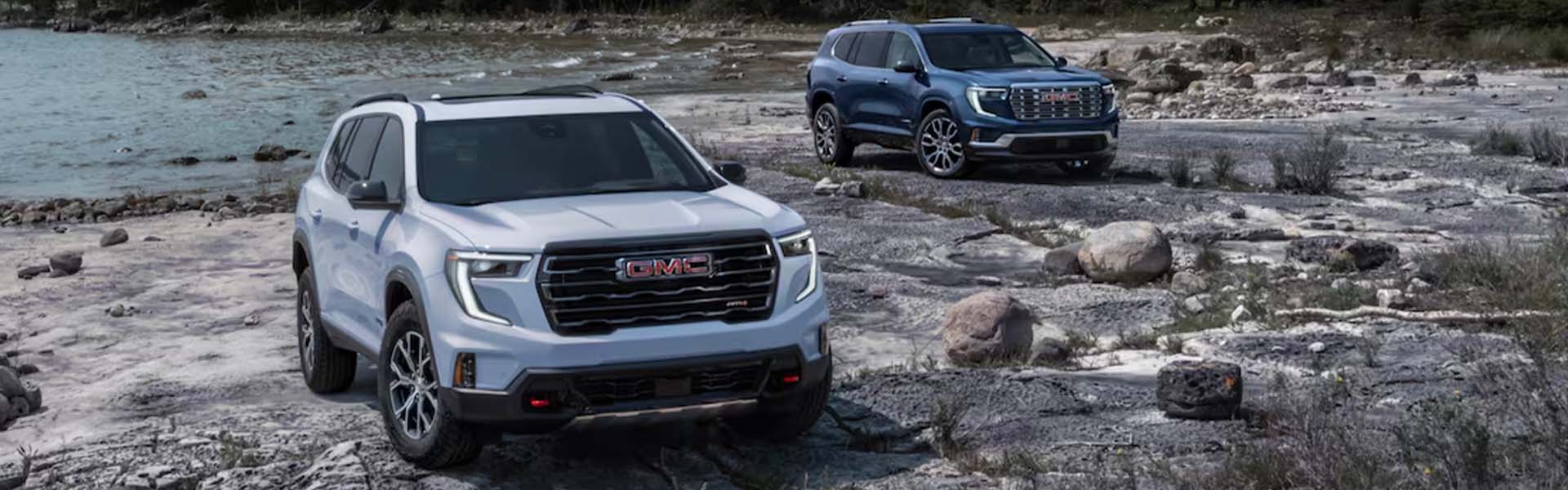 A white and blue Acadia is parked on a rocky riverbed with no water, surrounded by stones and natural scenery.
